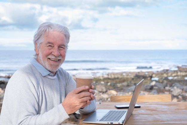 Sorrindo lindo homem sênior segurando a xícara de café ao ar livre sentado na mesa de madeira usando laptop