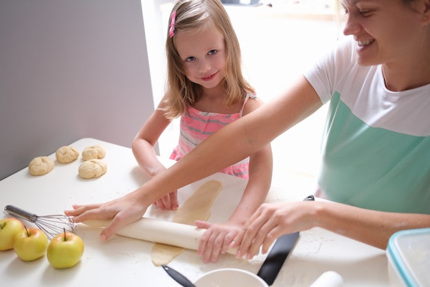 Sorrindo, mãe e filha estendem a massa com o rolo. Cozinhar alimentos doces junto com o conceito de crianças