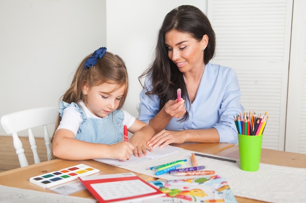 Sorrindo, mãe e filha se preparando para as aulas e desenha à mesa com lápis e tintas