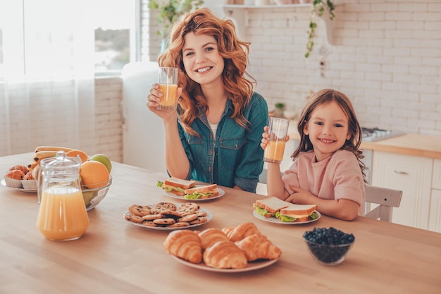 Sorrindo, mãe e filha segurando copos de suco de laranja e olhando para a câmera sentada na mesa da cozinha