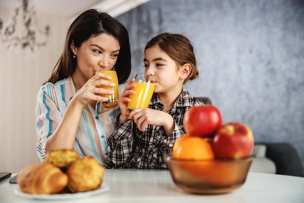 Sorrindo, mãe e filha sentada à mesa de jantar e tomando café da manhã saudável. eles estão bebendo suco de laranja.