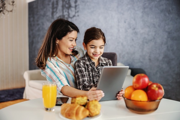 Sorrindo, mãe e filha sentada à mesa de jantar pela manhã e usando o tablet. Família feliz assistindo desenhos animados no tablet.