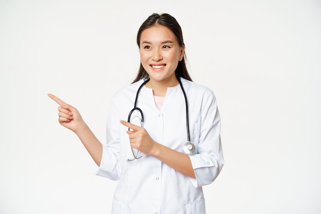 Sorrindo médica asiática em uniforme médico, apontando os dedos e olhando para a esquerda no anúncio co ...