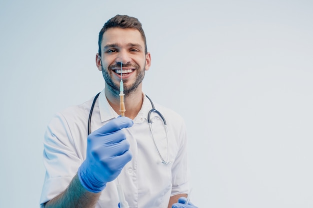 Sorrindo médico masculino europeu mostrando a seringa. Jovem barbudo com estetoscópio vestindo jaleco branco com luvas de látex. Isolado em um fundo cinza com luz turquesa. Sessão de estúdio. Copie o espaço.