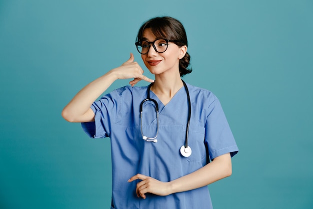 Sorrindo mostrando gesto de telefonema jovem médica usando estetoscópio uniforme de quinto isolado em fundo azul