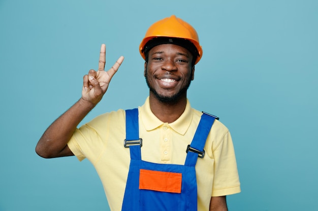 Sorrindo mostrando o gesto de paz jovem construtor afro-americano de uniforme isolado em fundo azul