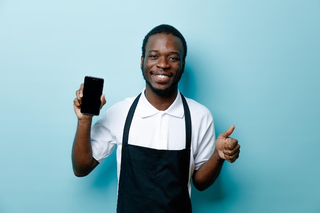 Sorrindo mostrando os polegares para cima jovem barbeiro americano africano de uniforme segurando o telefone isolado no fundo azul