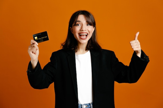 Sorrindo mostrando os polegares para cima segurando o cartão jovem linda mulher vestindo jaqueta preta isolada em fundo laranja