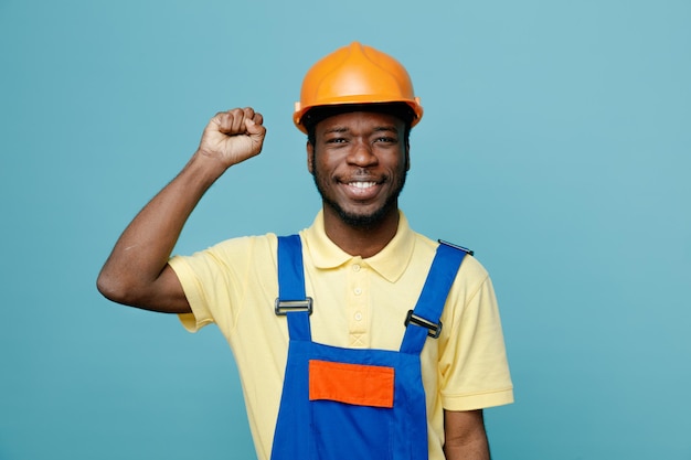 Sorrindo mostrando punho jovem construtor americano africano de uniforme isolado em fundo azul