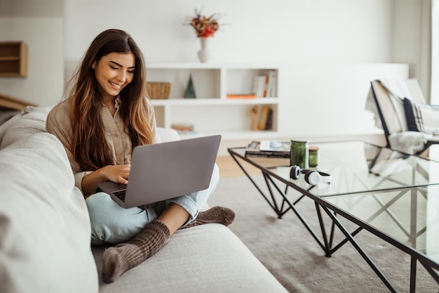 Sorrindo muito jovem mulher asiática digitando no laptop no interior da sala de estar