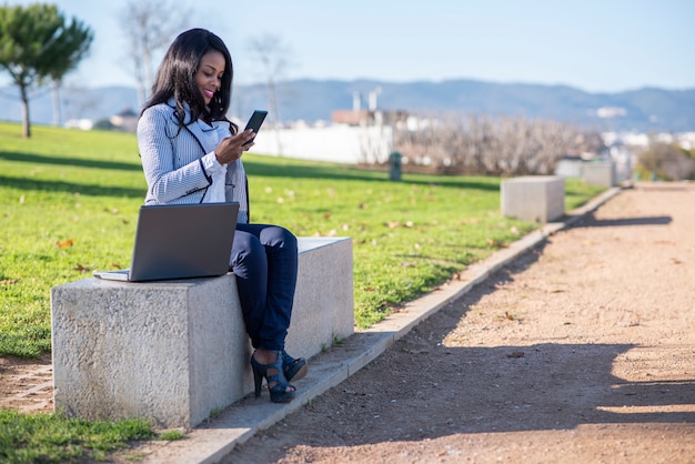 Sorrindo mulher afro-americana em um banco usando um laptop e telefone celular em um parque ao ar livre