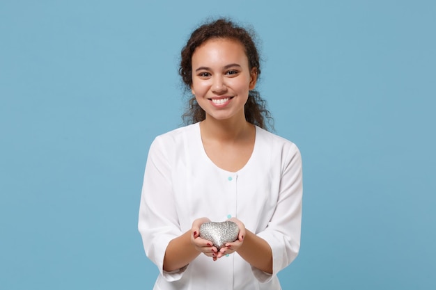 Sorrindo mulher afro-americana médica isolada no fundo da parede azul. Médica em vestido médico branco segura coração argent prateado. Conceito de saúde de medicina pessoal de saúde. Simule o espaço da cópia.