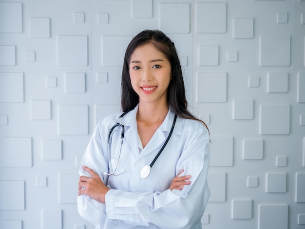 Sorrindo, mulher asiática bonita, retrato de cabelo comprido, de pé com os braços cruzados no consultório médico branco no hospital Confiante jovem praticante asiática com estetoscópio olhando para a câmera