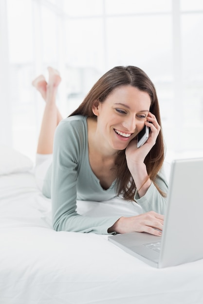 Sorrindo mulher casual usando celular e laptop na cama