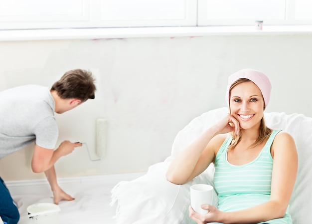 Sorrindo mulher caucasiana relaxando em um sofá enquanto namorado pintando o quarto