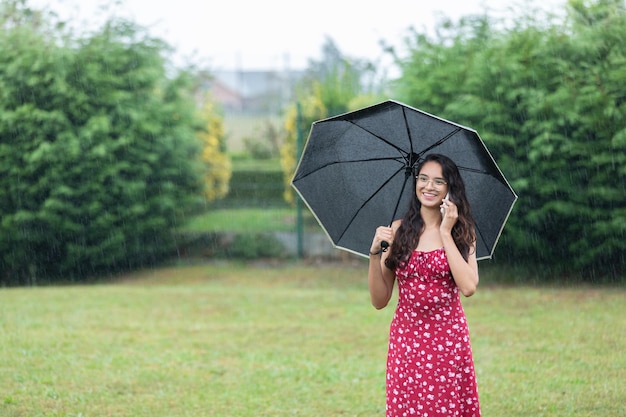 Sorrindo mulher étnica com guarda-chuva tendo telefonema no parque em dia chuvoso