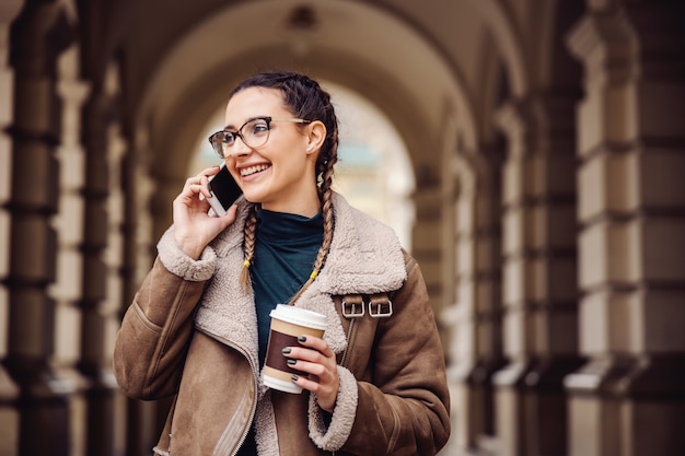 Sorrindo na moda universitária em pé no campus, segurando o copo descartável com café e conversando ao telefone.