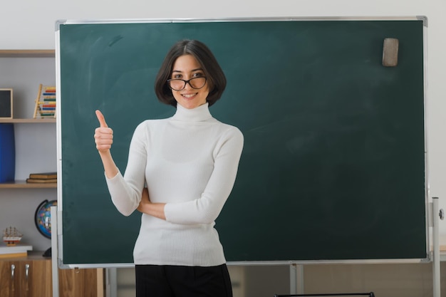 sorrindo olhando para a câmera mostrando os polegares para cima jovem professora usando óculos em pé na frente do quadro-negro em sala de aula