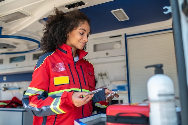 Foto sorrindo paramédico em um uniforme vermelho segurando embalagens de medicamentos em suas mãos no veículo ems