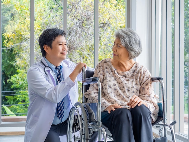 Sorrindo positivo médico homem asiático de jaleco branco fala com cuidado para paciente idosa idosa que está sentada em uma cadeira de rodas no consultório médico no hospital Cuidados de saúde e conceito médico