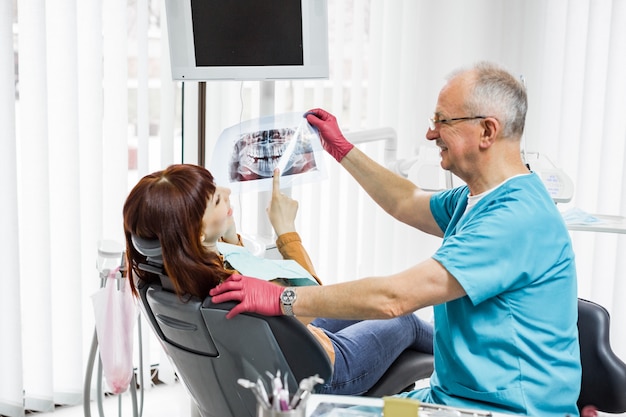 Sorrindo profissional dentista masculino sênior, mostrando o raio-x dental para paciente jovem, sentado na cadeira odontológica na clínica odontológica moderna