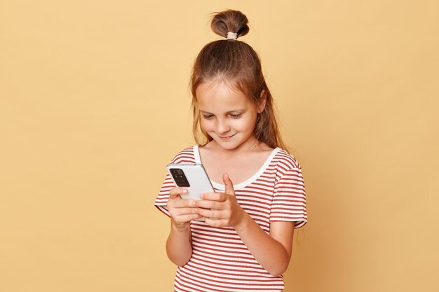 Foto sorrindo satisfeita menina de cabelos escuros vestindo camiseta listrada isolada sobre fundo bege usando smartphone jogando videogames navegando na internet