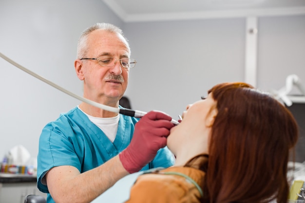 Sorrindo sênior experiente dentista homem realizando tratamento dentário de pacientes dente. jovem mulher na cadeira do dentista durante um procedimento odontológico.