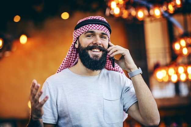 Sorrindo tatuado barbudo na moda muçulmano sentado no café pela manhã e tendo uma conversa de camponês com seu amigo no celular.