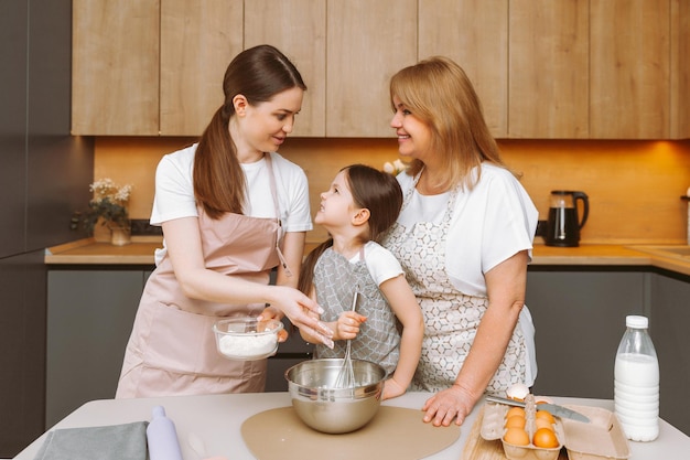 Sorrindo, três gerações de mulheres se divertem fazendo massa na cozinha Uma garotinha feliz com a mãe e a avó mais velha estão preparando bolos ou biscoitos