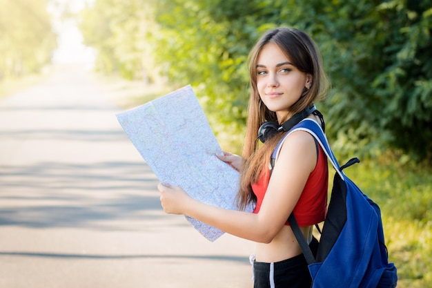 Sorrindo turista feminina olhando para trás, segurando um mapa e mochila pesada enquanto fazia uma viagem para o campo