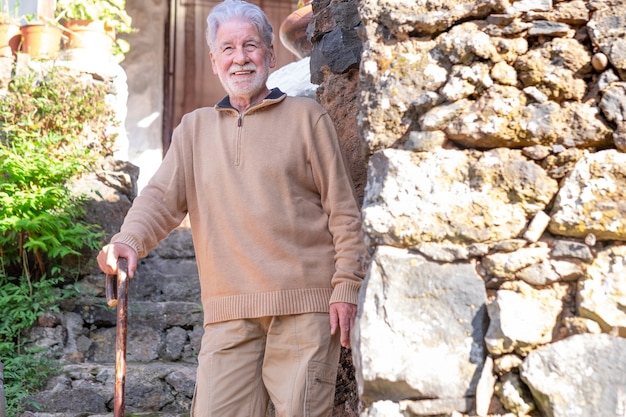 Sorrindo velho homem sênior com camisola de inverno ao ar livre de uma casa rural apoiada na bengala. homem caucasiano com barba e cabelo branco