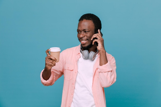 Sorrir fala ao telefone segurando uma xícara de café jovem afro-americano usando fones de ouvido no pescoço isolado em fundo azul