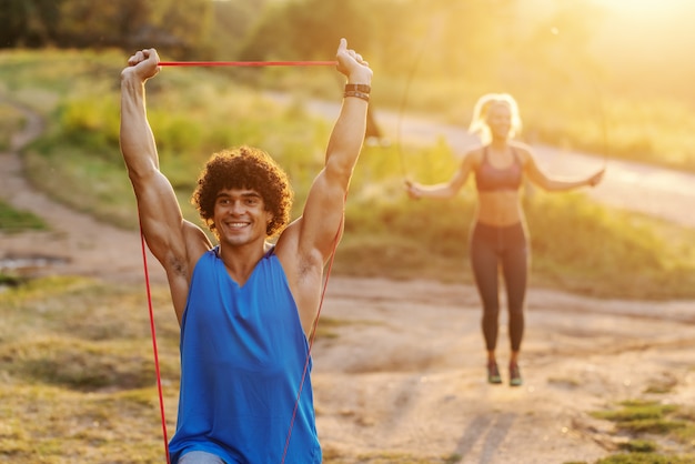 Foto sorrir homem desportivo caucasiano com cabelos cacheados, fazendo exercícios de alongamento com corda na natureza em dia ensolarado de verão. no fundo mulher pulando corda.