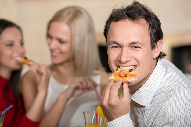 Sorrir homem está comendo uma pizza. Duas garotas estão conversando.