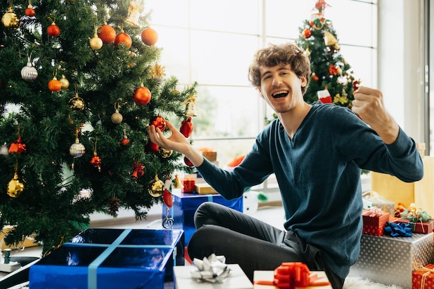 Sorrir jovem bonito sentar no chão gosta de decorar a árvore de Natal em casa. Comemorando o Ano Novo sozinho.