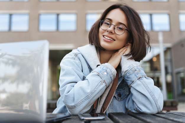 Sorrir uma jovem sonhadora de óculos, olhando para a câmera feliz, relaxante tendo uma conversa com o colega durante a pausa para o almoço no campus da universidade, usando o laptop, preparar o ensaio.