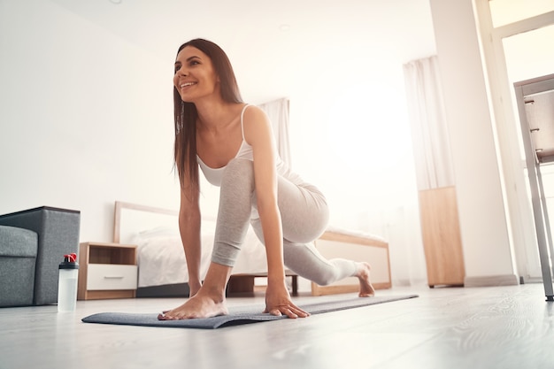 Sorriso amigável. Mulher alegre usando tapete de ioga enquanto faz exercícios de alongamento