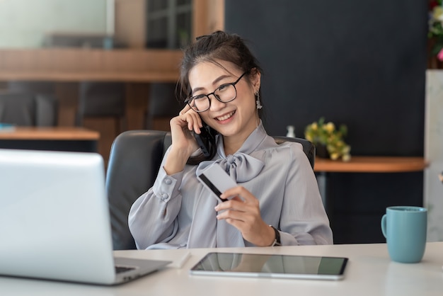 Sorriso bonito mulher asiática falando no celular feliz segurando cartão de crédito no escritório.