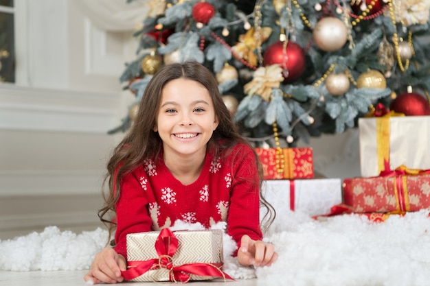 Sorriso de criança feliz com caixa de presente deitada na neve artificial na árvore de Natal, ano novo.