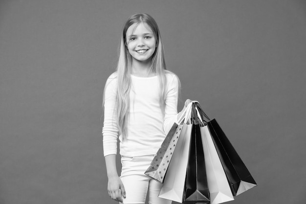 Sorriso de menina com sacolas de compras em fundo violeta criança feliz com sacos de papel em fundo roxo comprador de criança em roupas casuais férias e celebração compras e venda na sexta-feira negra