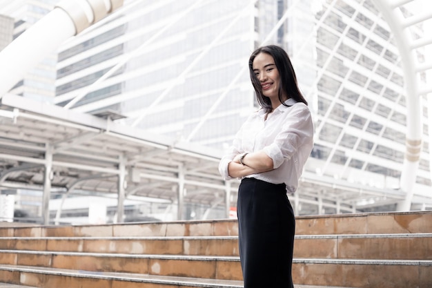 Sorriso do retrato Menina asiática do trabalhador de escritório com os braços cruzados no espaço ao ar livre e da cópia. Sorrindo feliz empresária tailandesa com fundo da cidade.