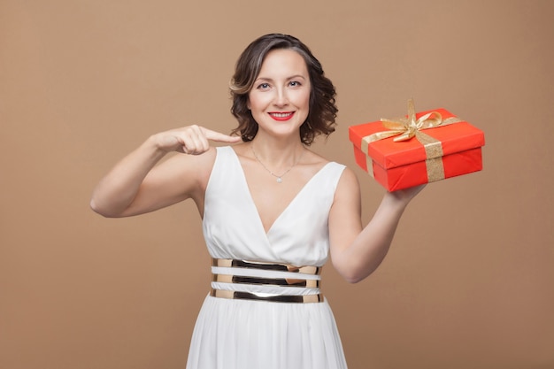 Sorriso elegante mulher morena com vestido branco, apontando para a caixa de presente vermelha e olhando para a câmera com uma cara feliz. conceito de emoção e sentimento. foto de estúdio, interna, isolada em fundo marrom claro