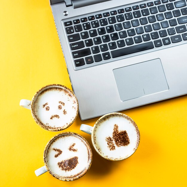 Sorriso emoji pintado em copos de cappuccino ao lado do laptop em um fundo amarelo