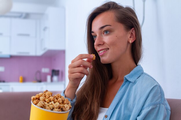 Sorriso feliz jovem mulher descansando e comendo pipoca de caramelo crocante durante assistir filme em casa