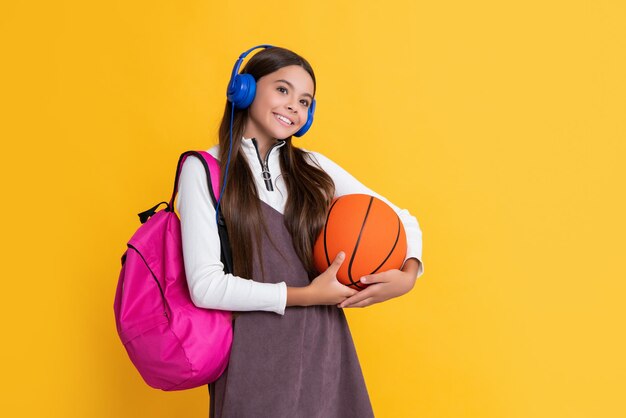 Sorriso infantil em fones de ouvido com mochila escolar e bola de basquete em fundo amarelo