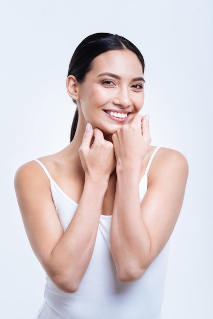 Sorriso radiante. Linda jovem de cabelos escuros em um top branco posando contra uma parede branca enquanto ri