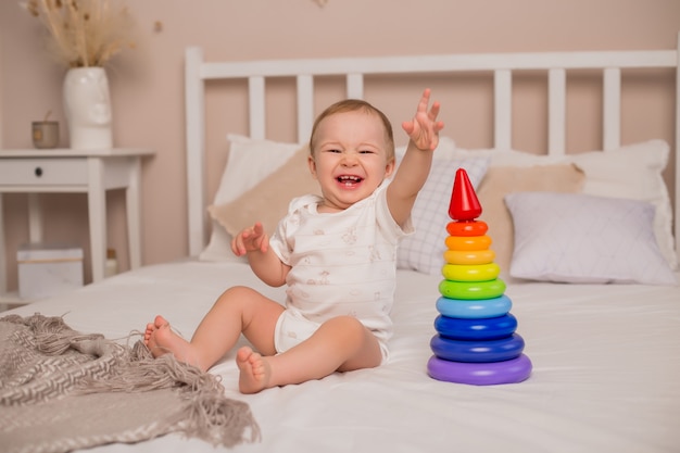 Sorrisos de menino bonito da criança sentada na cama com pirâmide multicolorida