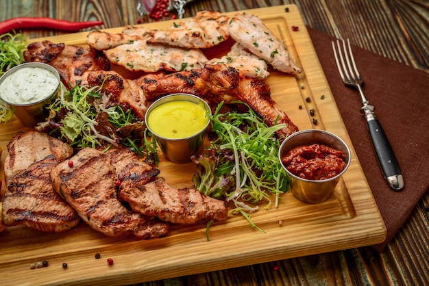 Sortiertes köstliches gegrilltes Fleisch und Gemüse mit frischem Salat und Bbq-Soße auf Schneidebrett auf hölzernem Hintergrund. Große Reihe von warmen Fleischgerichten