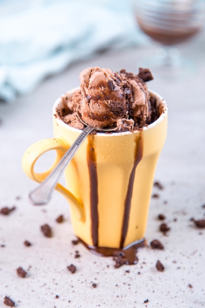 Foto sorvete castanho chocolate em caneca e colher amarela regular, em azul, com cobertura e migalhas.