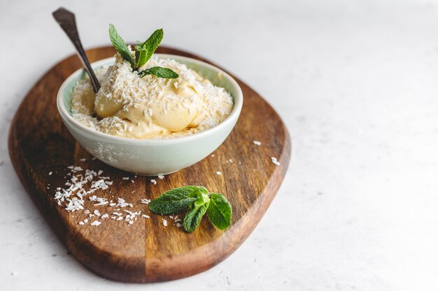 Sorvete vegano caseiro em uma tigela é feito de manga congelada e leite de coco servido por hortelã fresca e flocos em uma placa texturizada de madeira